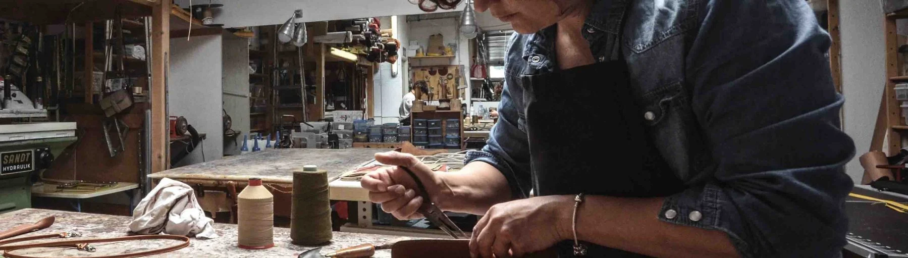 Une personne portant un tablier travaille sur une pièce de cuir dans un atelier rempli d’outils et de matériaux.