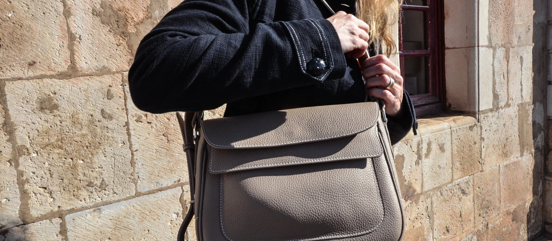 Personne en manteau noir tenant un sac à main beige devant un mur de pierre.