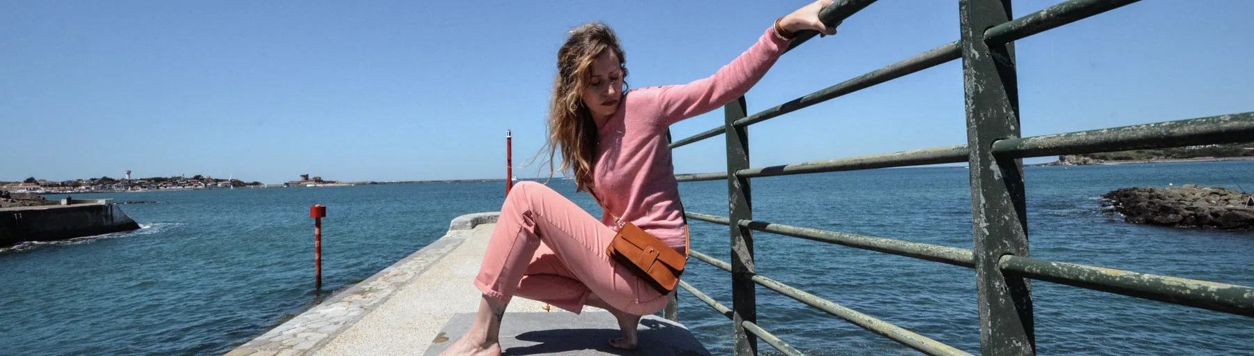 Une femme en tenue rose et sac orange est assise sur la balustrade d'un quai au bord de la mer par temps clair.