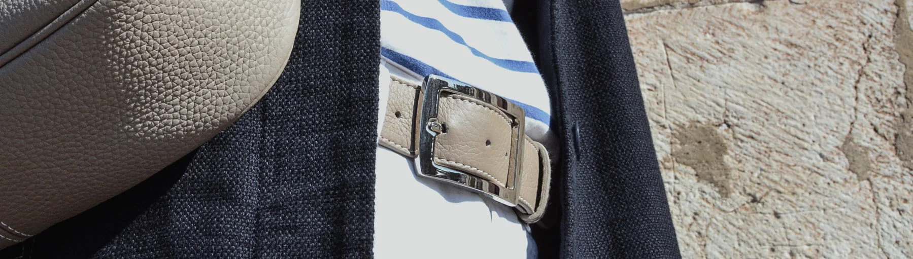 Gros plan d'une personne portant une veste noire, une chemise rayée et un sac ceinture beige avec une finition texturée, debout contre un mur de pierre.