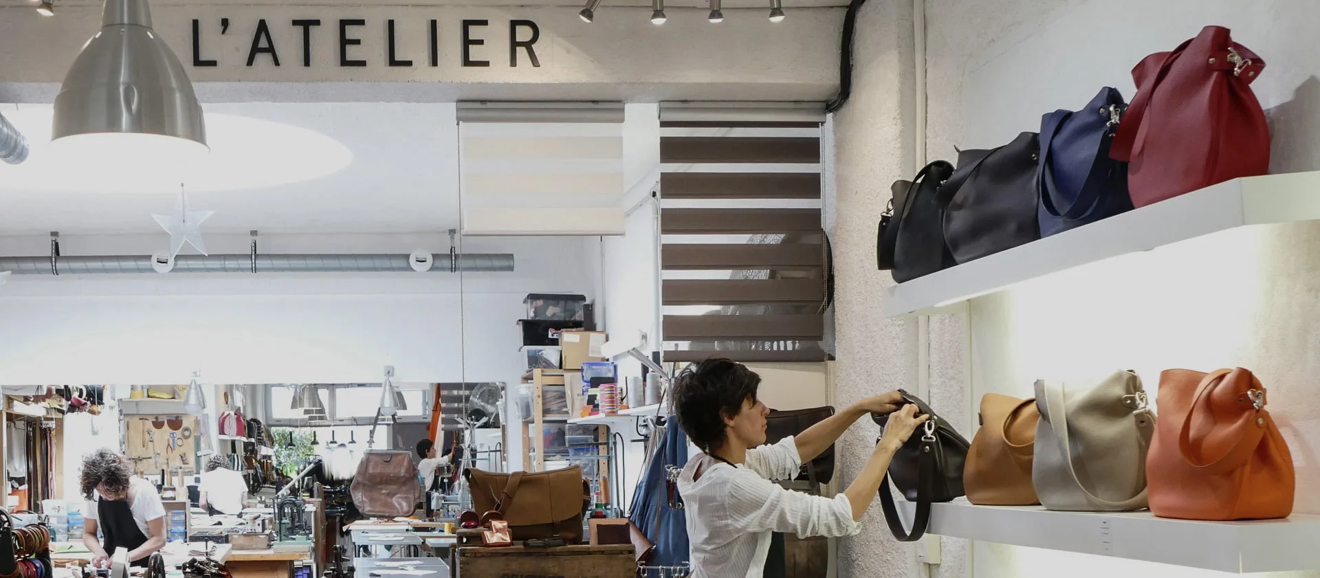 Une personne range des sacs à main sur une étagère dans un magasin étiqueté LATELIER, avec des sacs de différentes couleurs exposés. Une autre personne travaille à une table en arrière-plan.