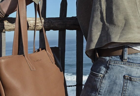 Une personne portant un jean et une chemise verte se tient près d'une clôture en bois au bord de la mer, avec un sac fourre-tout en cuir marron suspendu à son épaule.