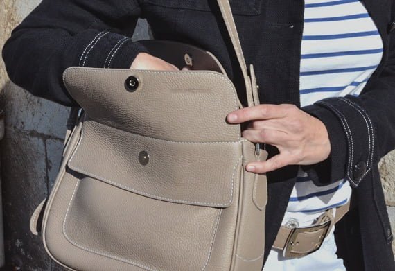Une personne portant une chemise rayée et une veste noire ouvre un sac à main en cuir beige.