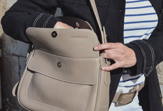 Une personne portant une chemise rayée et une veste noire ouvre un sac à main en cuir beige.