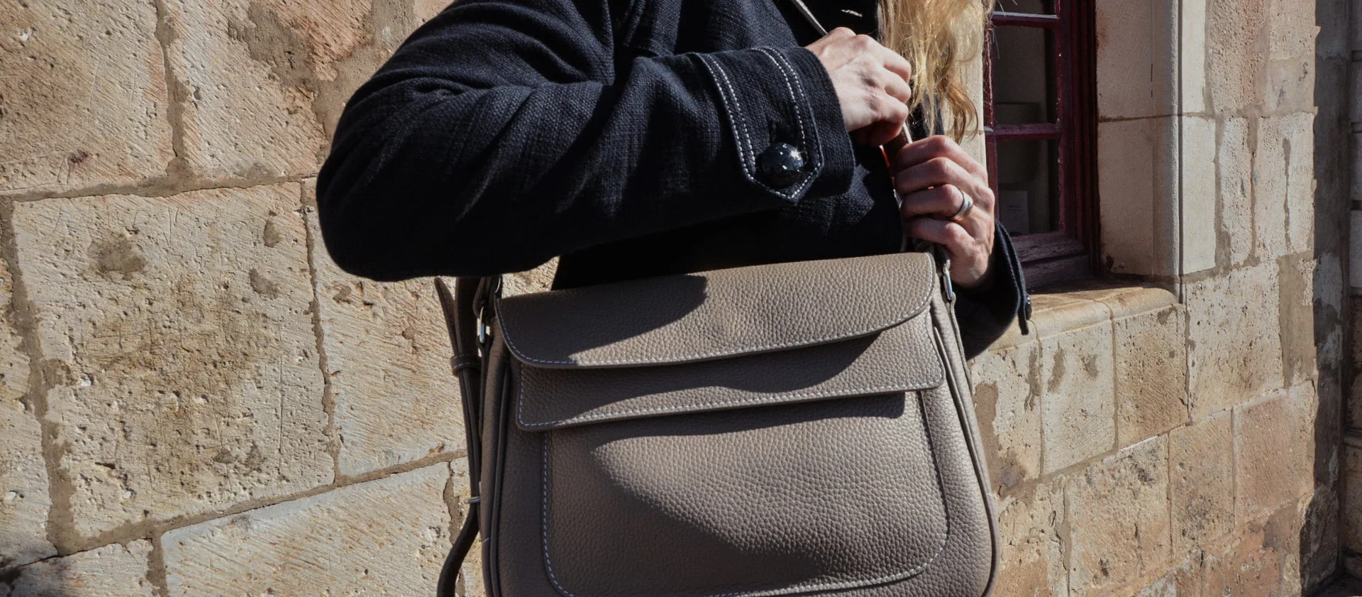 Personne tenant un sac à main en cuir taupe, debout contre un mur de pierre avec une fenêtre.