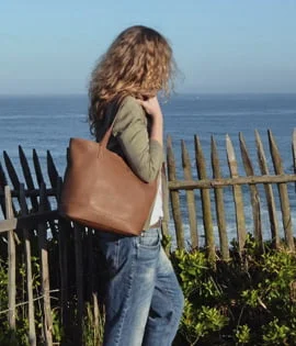 Une personne aux cheveux bouclés se tient près d'une clôture en bois près de l'océan, portant un sac marron et portant un jean et une veste.