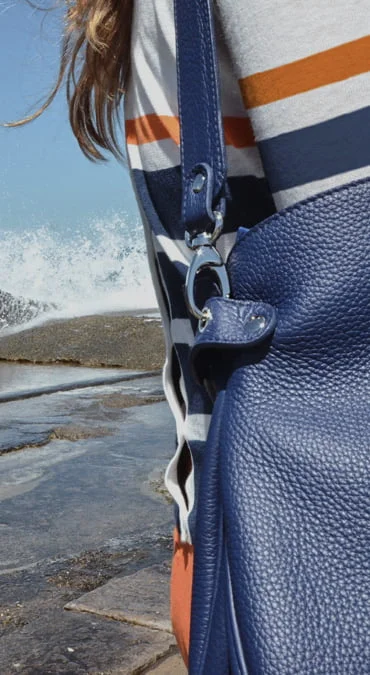 Personne au bord de l'océan portant une chemise rayée et portant un sac en cuir bleu avec un fermoir en métal, des vagues éclaboussant en arrière-plan.