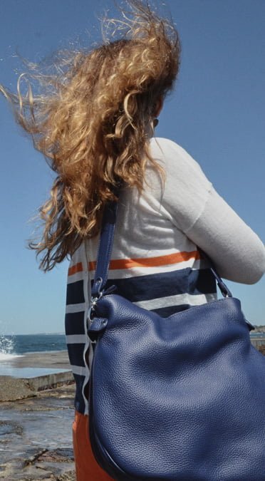Une personne aux cheveux ondulés et au pull rayé tient un grand sac bleu, face à l'océan par une journée venteuse.