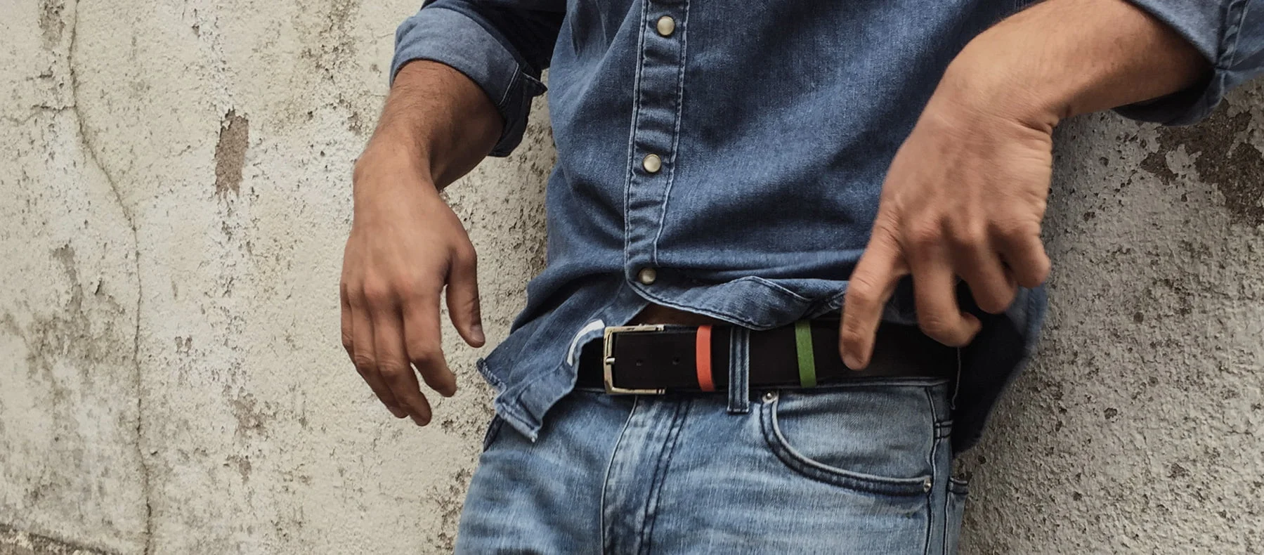 Une personne portant une chemise en jean et un jean se tient debout contre un mur texturé, ajustant une ceinture noire.
