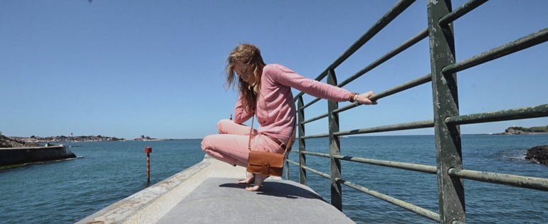 Une personne vêtue de rose est accroupie sur un rebord de béton au bord de l'océan, s'accrochant à une balustrade en métal.