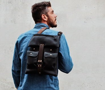 Une personne portant une veste en jean porte un sac à dos en cuir noir avec des bretelles marron sur un fond uni et clair.