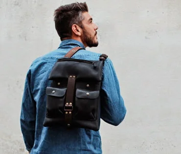 Une personne portant une veste en jean porte un sac à dos en cuir noir avec des bretelles marron sur un fond uni et clair.
