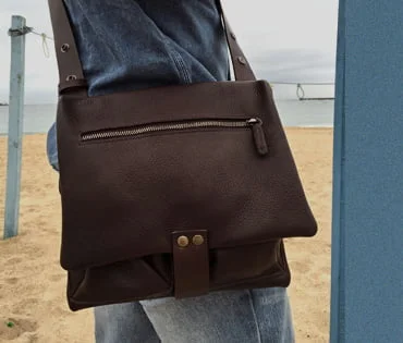 Personne tenant un sac en cuir foncé avec une fermeture éclair sur un fond de plage de sable.