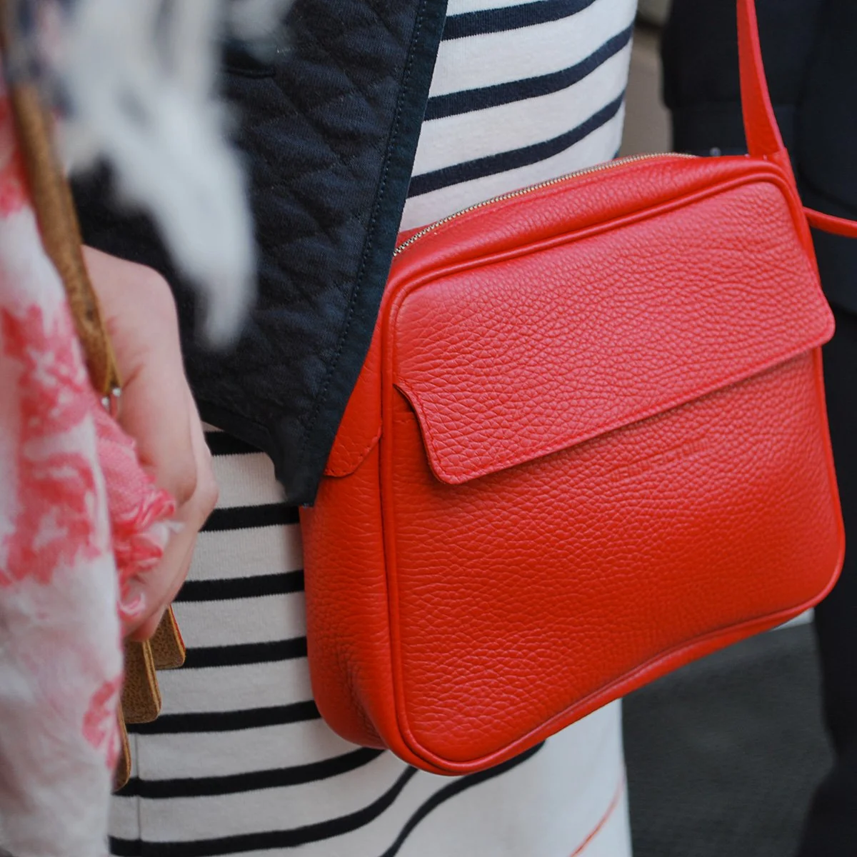 Une personne tient un petit sac à bandoulière en cuir orange avec un rabat, portant une tenue rayée noire et blanche.