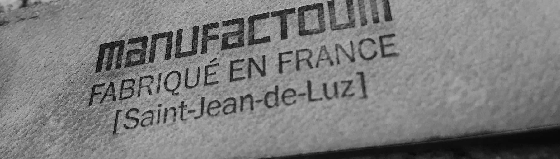 Gros plan d'une étiquette portant le texte MANUFACTOUM FABRIQUÉ EN FRANCE [Saint-Jean-de-Luz] en noir sur une surface texturée.