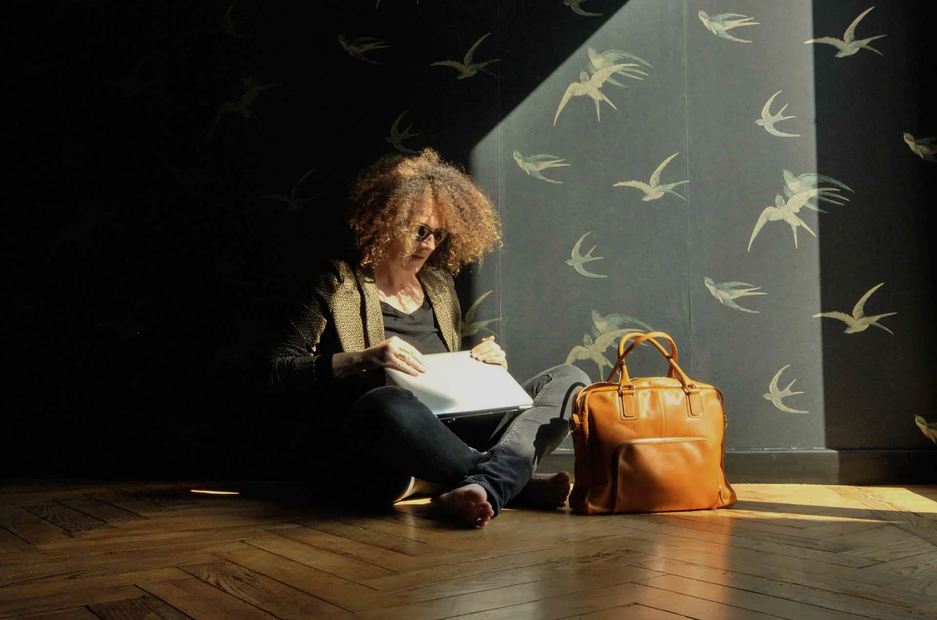 Une personne aux cheveux bouclés est assise sur un plancher en bois et lit un livre. À côté d'elle se trouve un sac en cuir marron. Le mur est orné de motifs d'oiseaux et un faisceau de lumière illumine la scène.