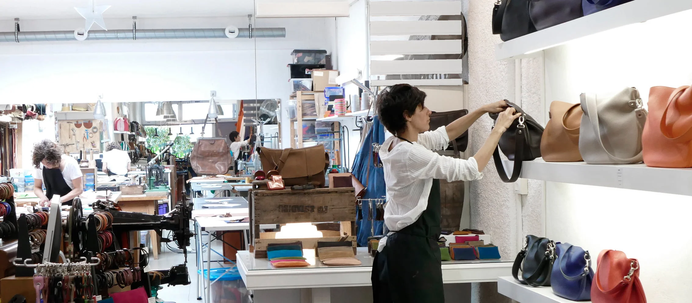 Deux personnes travaillent dans un magasin de maroquinerie, l'une dispose des sacs à main sur une étagère et l'autre travaille en arrière-plan près d'une table avec divers matériaux.