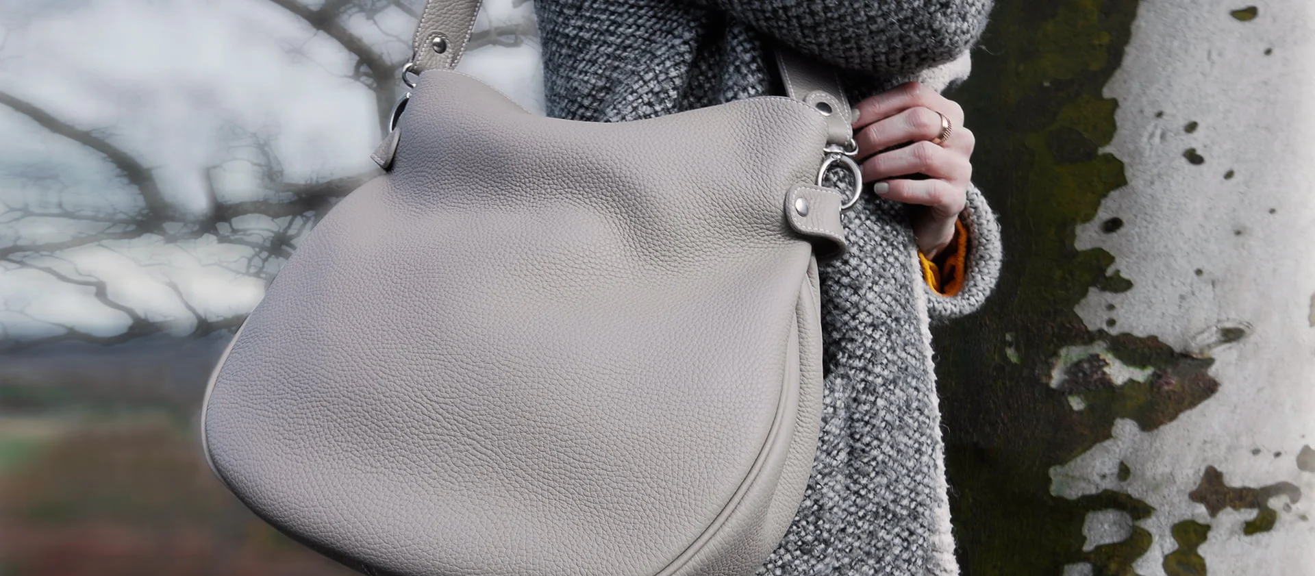 Personne portant un manteau gris tenant un grand sac à main gris texturé à l'extérieur près d'un arbre.