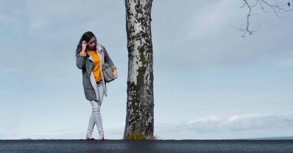 Une femme portant un manteau gris et un haut jaune se tient pieds nus sur une route à côté d'un arbre contre un ciel nuageux.