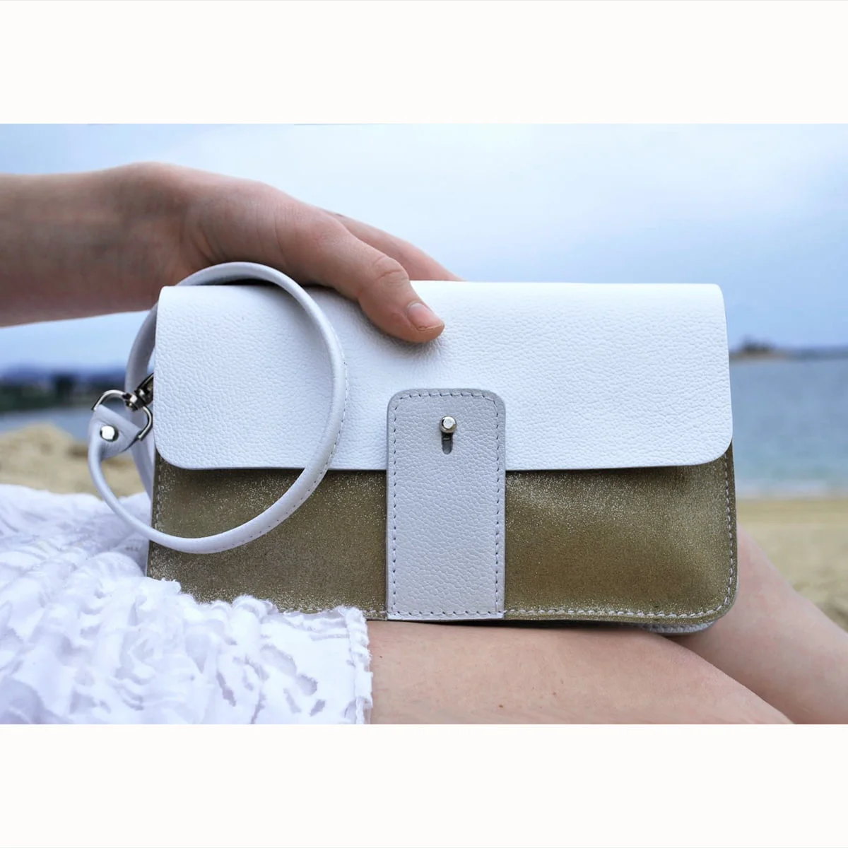 Une main tient une pochette blanche et dorée avec une dragonne, sur fond de plage et d'océan.