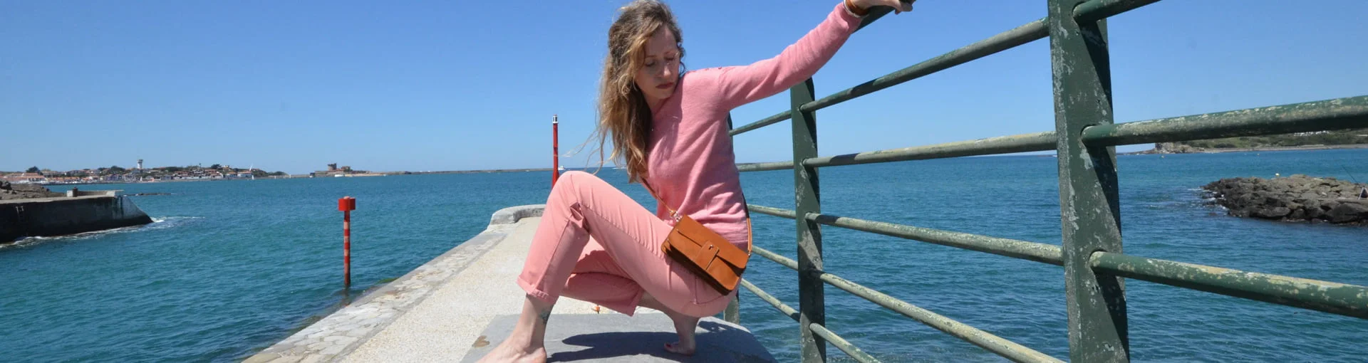 Une personne vêtue d'une tenue rose est accroupie sur une jetée, s'accrochant à une balustrade en métal verte avec vue sur l'océan par temps clair.