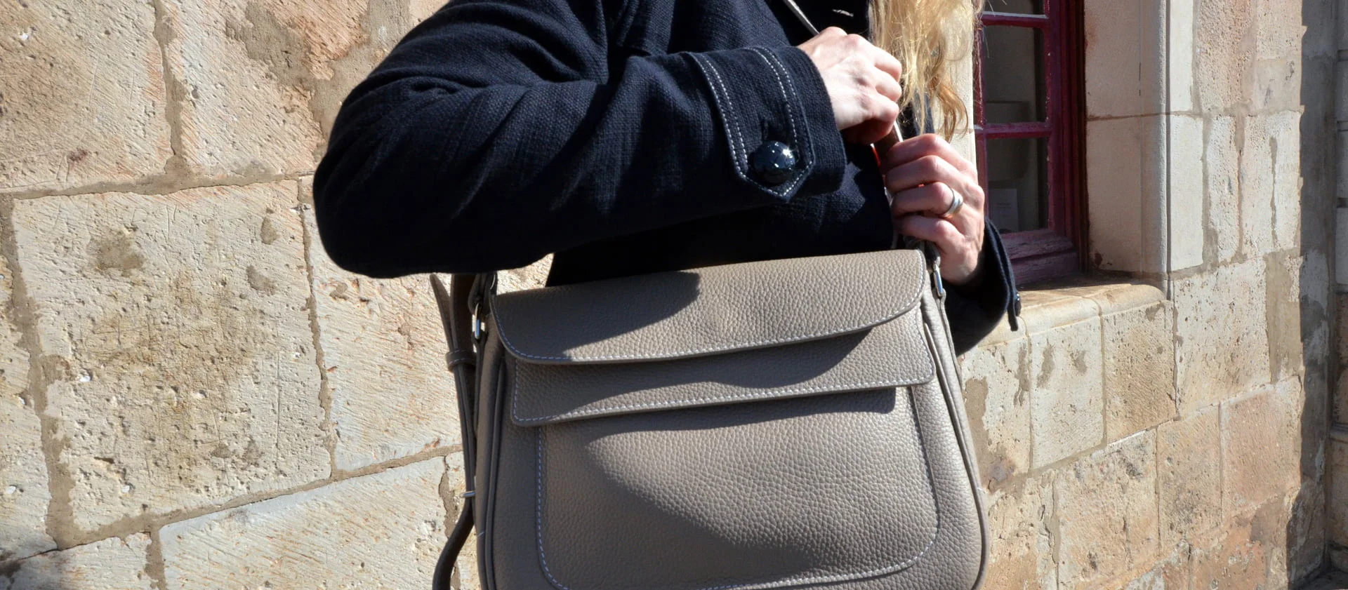 Personne tenant un sac à main en cuir beige tout en portant un manteau foncé, debout contre un mur de pierre avec une fenêtre.