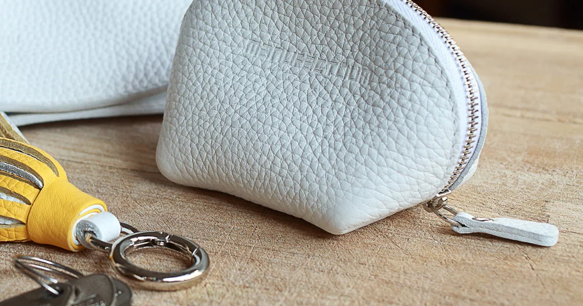 Un petit porte-monnaie en cuir texturé blanc avec une fermeture éclair est placé sur une surface en bois à côté d'un trousseau de clés avec un porte-clés en cuir jaune.