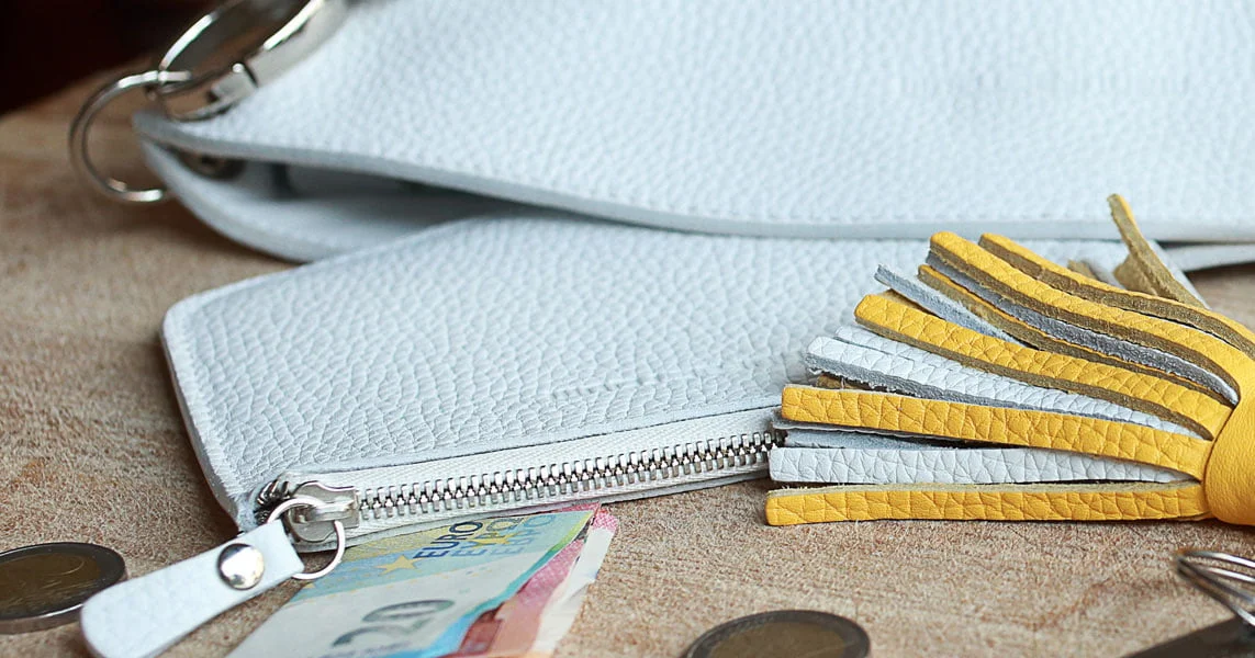 Un sac à main blanc avec fermeture éclair et pompon jaune est ouvert sur une table. À proximité se trouvent des billets et des pièces en euros.