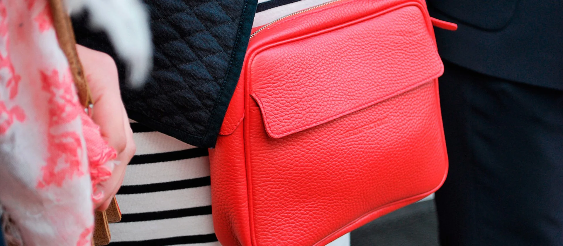 Gros plan d'une personne tenant un sac à main en cuir texturé rouge corail vif, avec un vêtement rayé noir et blanc partiellement visible.
