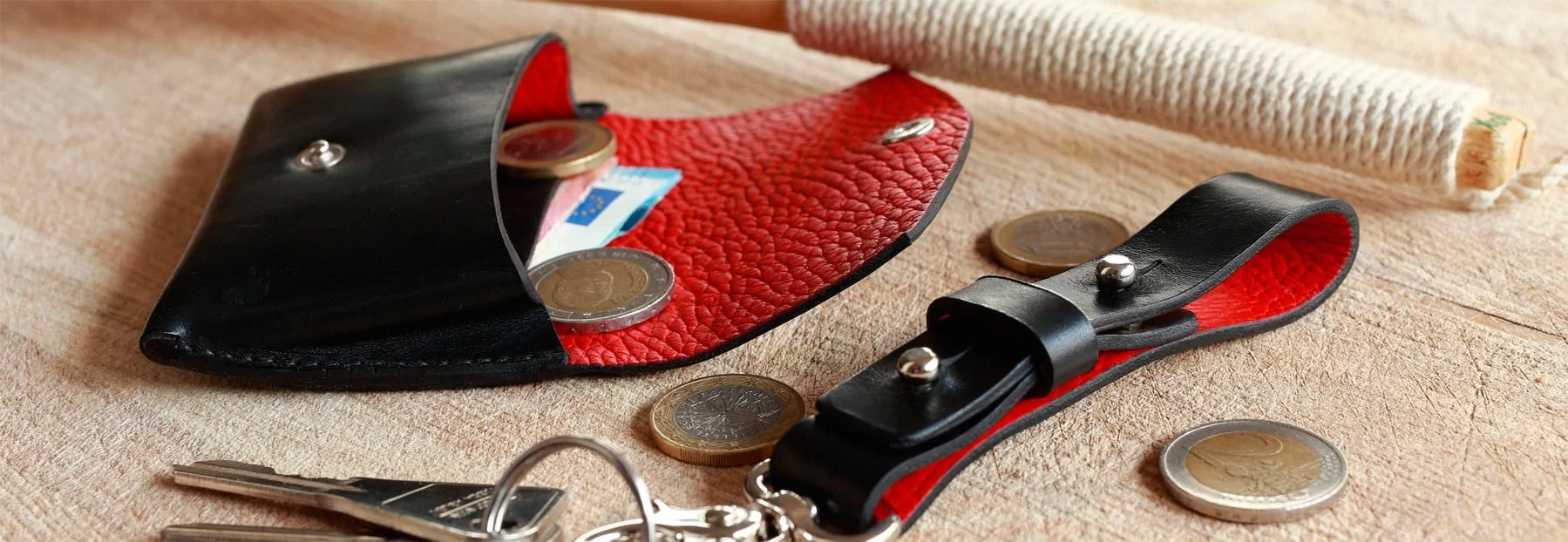 Un porte-monnaie en cuir noir et rouge est ouvert sur une surface en bois avec des pièces en euros éparpillées. Un trousseau de clés est attaché à un porte-clés en cuir noir assorti.