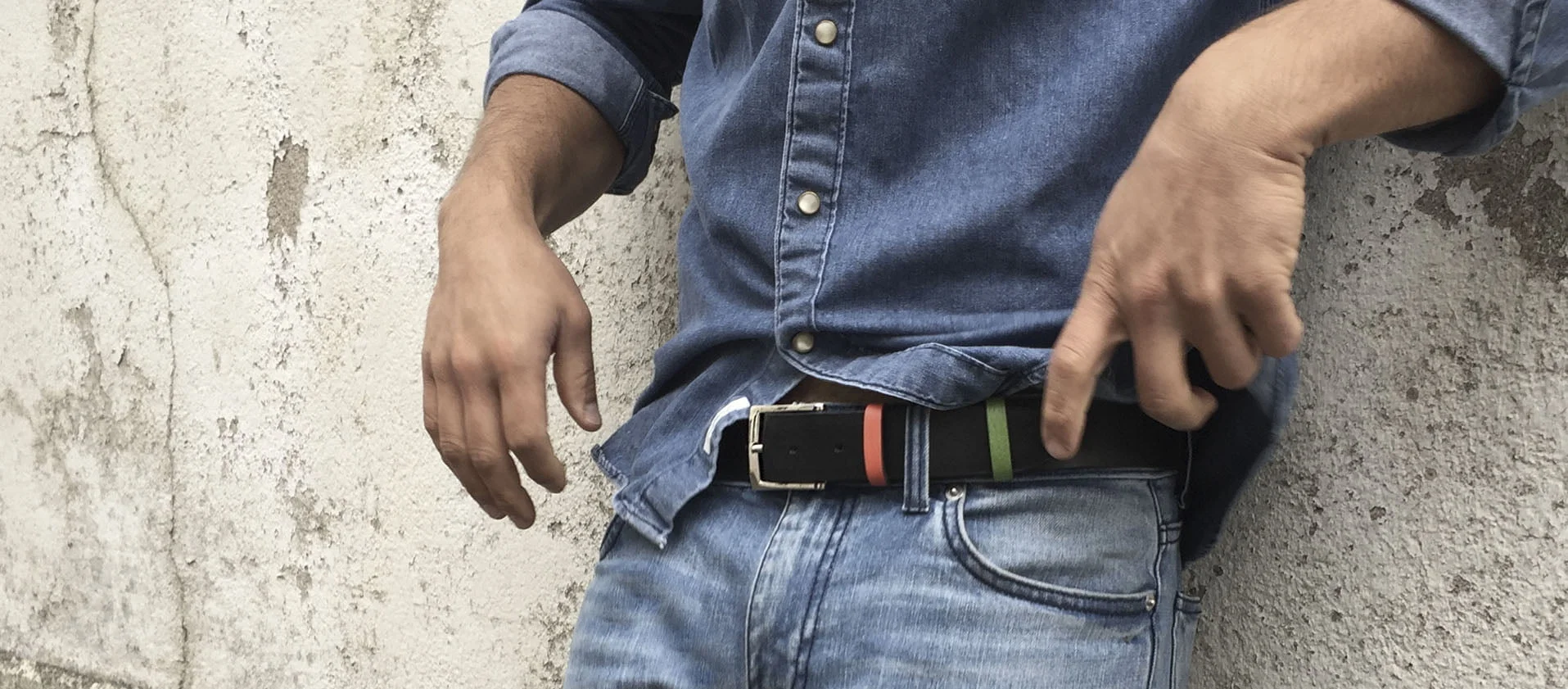 Une personne portant une chemise en jean bleu et un jean se tient debout contre un mur en béton, ajustant une ceinture colorée.