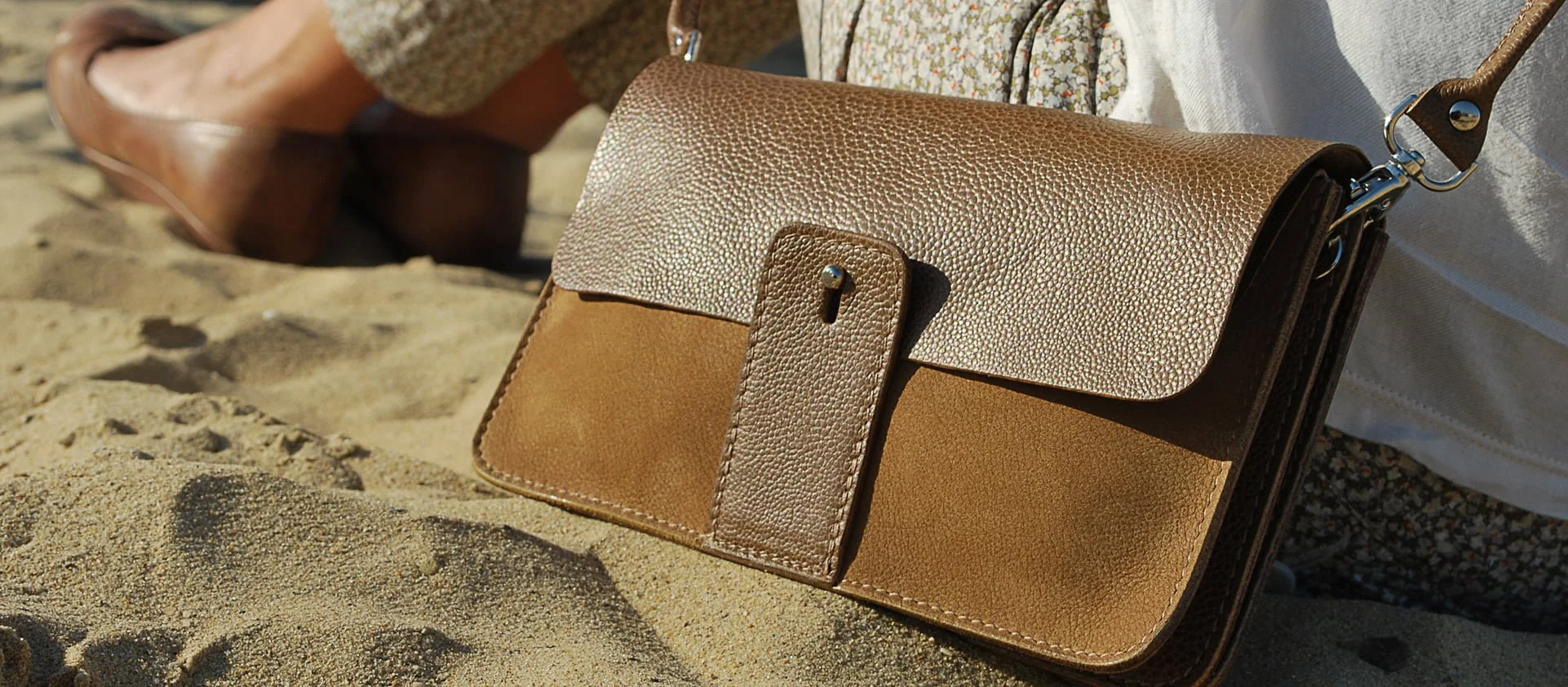 Sac à main en cuir marron posé sur le sable avec les jambes d'une personne en arrière-plan.