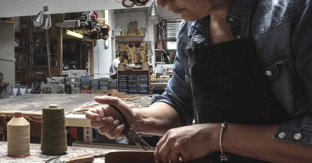 Une personne portant un tablier travaille avec des outils sur un établi encombré dans un atelier. Une autre personne est visible à l'arrière-plan au milieu des étagères et du matériel.