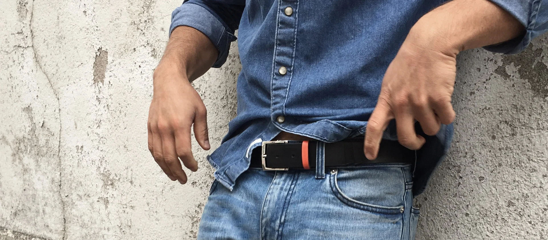 Personne portant une chemise en jean bleu et un jean, ajustant une ceinture noire, debout contre un mur texturé.