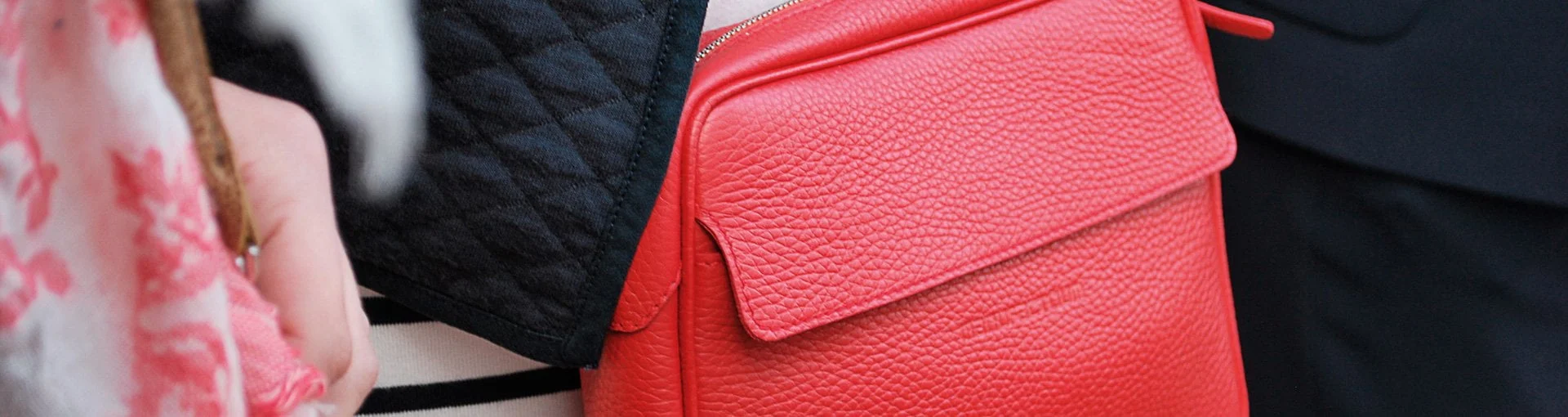 Gros plan d'une personne tenant un sac à main rouge texturé, à côté d'un sac matelassé noir. L'arrière-plan montre une partie d'un tissu à motifs floraux.