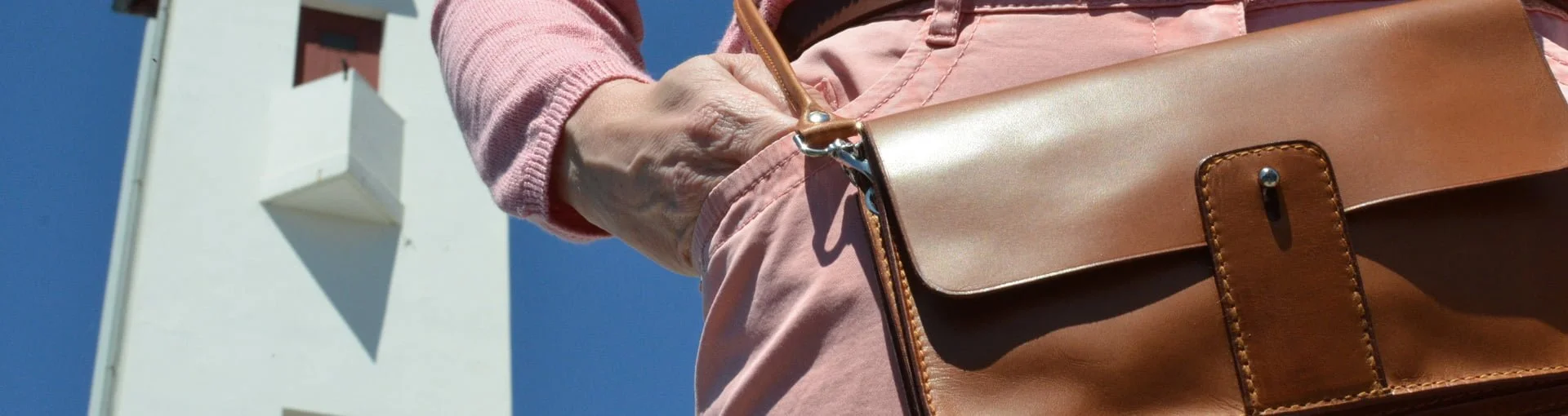 Personne tenant un sac en cuir marron, debout devant un bâtiment blanc contre un ciel bleu clair.
