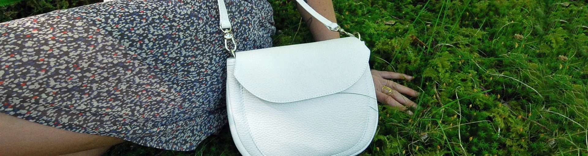Personne portant une robe à motifs allongée sur l'herbe avec un sac à bandoulière en cuir blanc posé sur le côté.