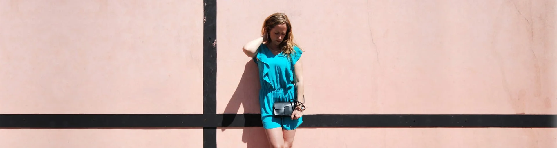 Une femme en robe bleue se tient contre un mur rose avec une bordure noire, regardant vers le bas.