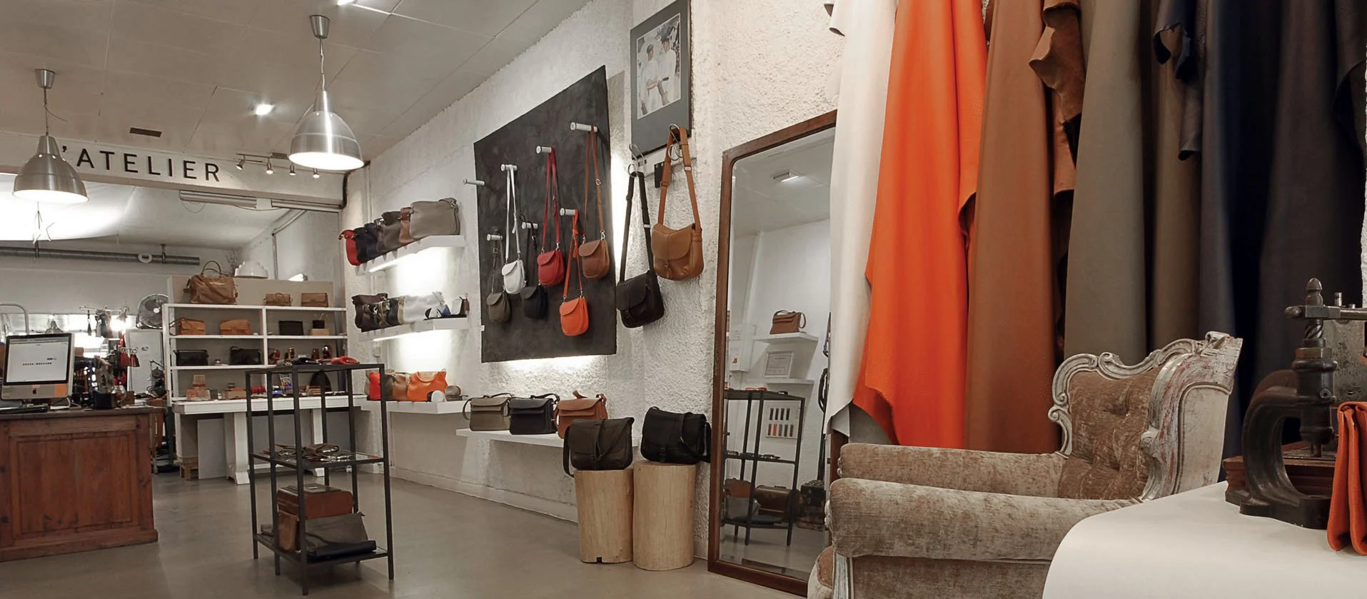 À l'intérieur d'une boutique avec des sacs à main sur des étagères, un grand miroir et une chaise vintage. Des rouleaux de tissu et des matériaux en cuir sont à l'arrière-plan.