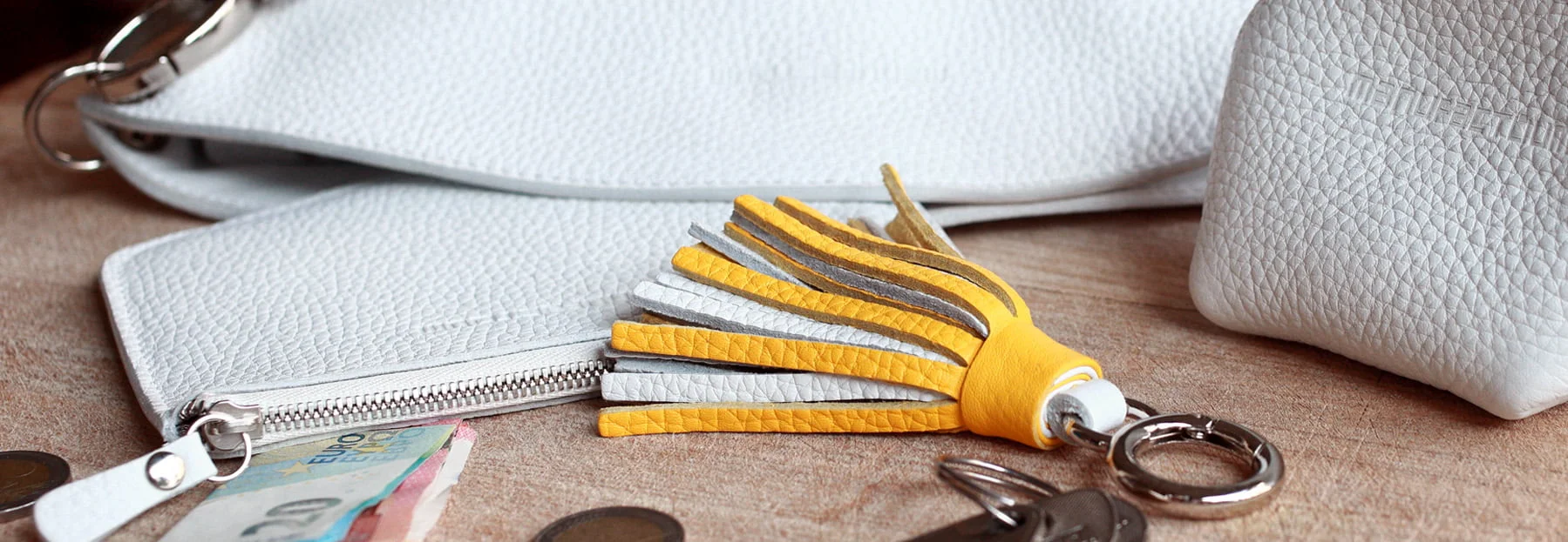 Des accessoires en cuir blanc, un porte-clés à pompon jaune et gris, des pièces de monnaie, des clés et un billet de 5 euros sont posés sur une surface en bois.