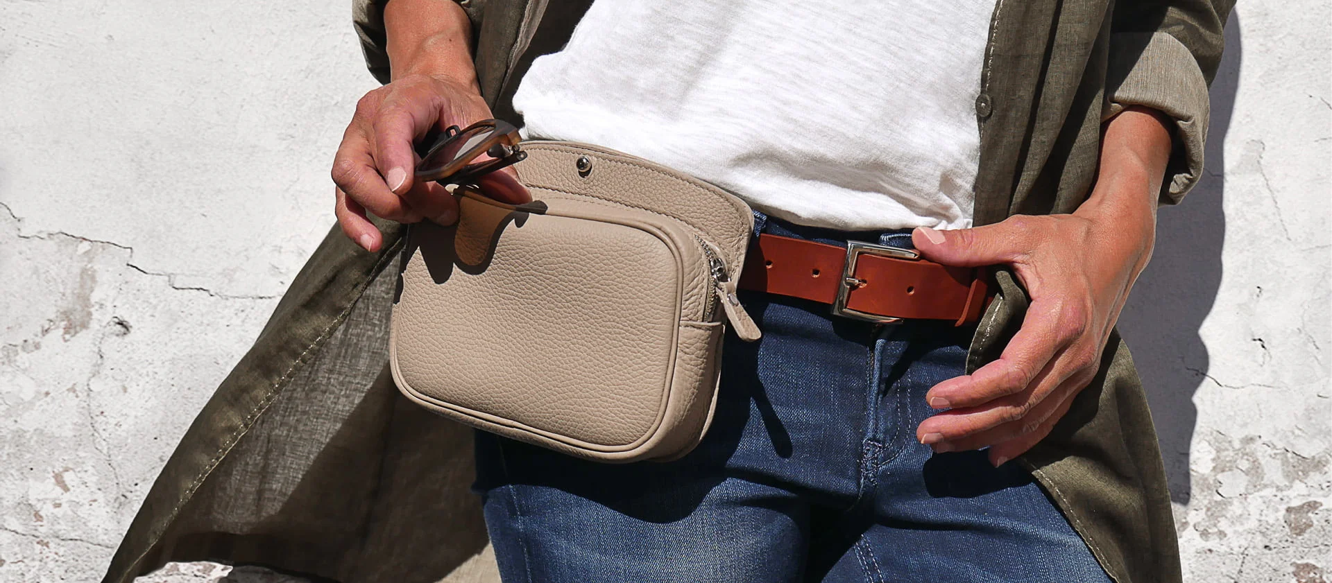 Une personne portant un manteau vert, une chemise blanche et un jean tient des lunettes de soleil et une pochette en cuir beige attachée à une ceinture marron.