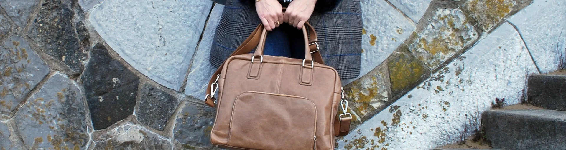 Personne tenant un sac à main en cuir marron devant un mur de pierre.