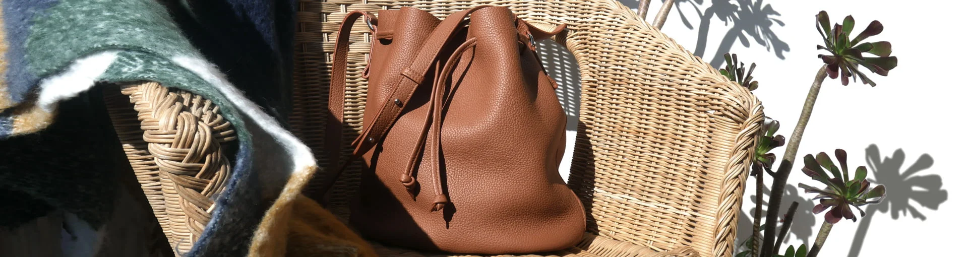 Un sac en cuir marron sur une chaise en osier à côté d'une plante et d'un tissu drapé au soleil.