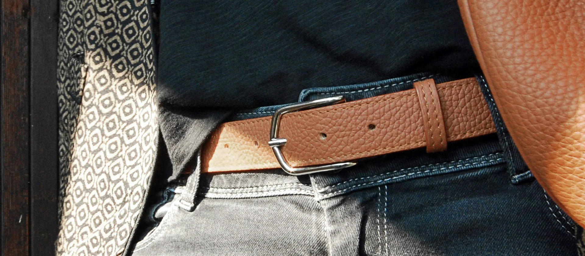 Ceinture en cuir beige avec boucle argentée portée avec un jean noir et une chemise foncée. Un tissu à motifs est en arrière-plan.