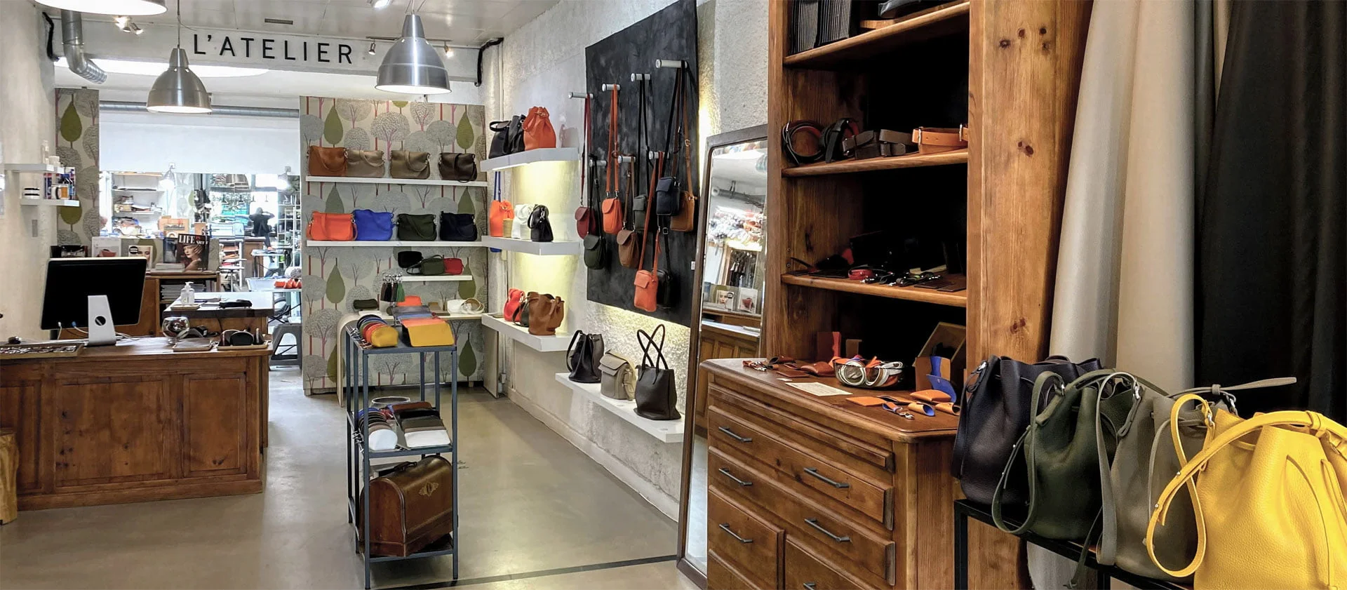 Une boutique avec des sacs en cuir exposés sur des étagères et une vitrine en bois. On voit un comptoir avec un ordinateur à gauche, et le magasin porte une enseigne indiquant Latelier.