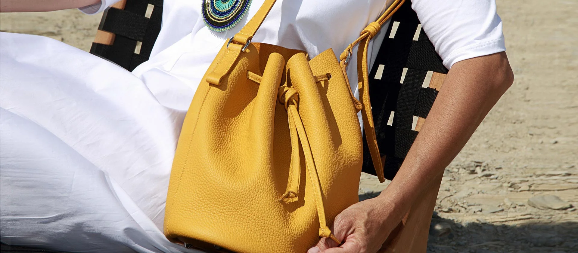 Personne tenant un sac à main jaune à cordon alors qu'elle est assise sur une plage, portant une tenue blanche avec un collier coloré.