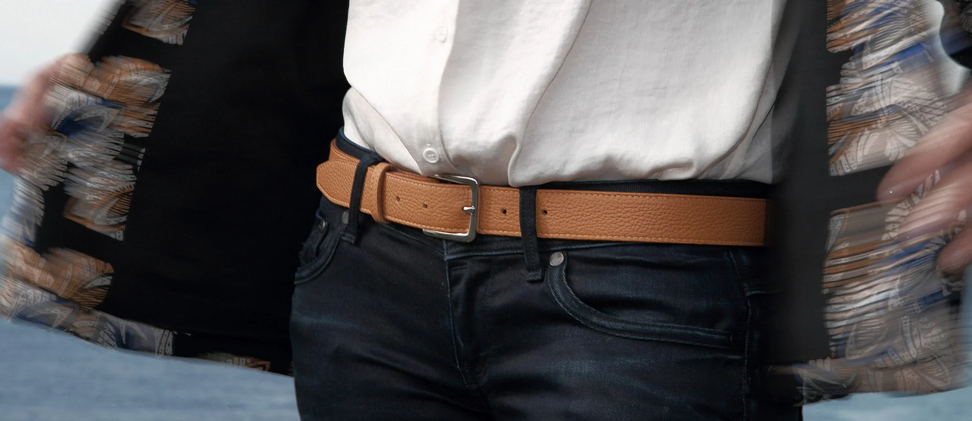 Personne portant une ceinture beige avec une boucle argentée, un jean foncé, une chemise blanche et un manteau ouvert à motifs.