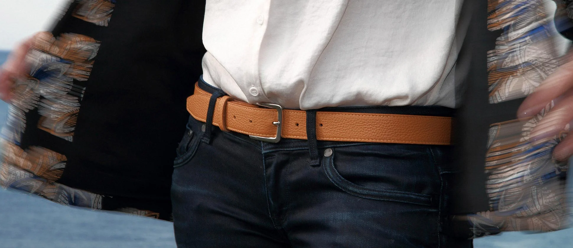 Une personne portant une chemise blanche, une veste noire à motifs et une ceinture beige avec un jean se tient à l'extérieur.