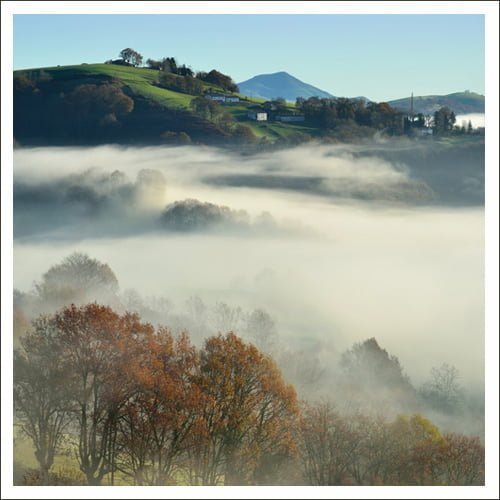 Un paysage brumeux avec des collines ondulantes, des arbres aux couleurs d'automne et des montagnes lointaines sous un ciel clair. Le brouillard recouvre les zones basses, obscurcissant partiellement la vue.