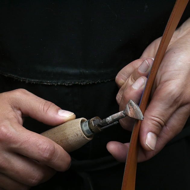 Gros plan de mains utilisant un outil pour travailler sur un bracelet en cuir sur un fond sombre.